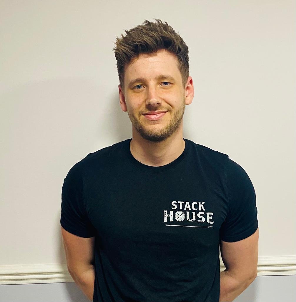 Person wearing a black "Stack House" t-shirt, smiling with arms behind back, standing against a light-colored wall.