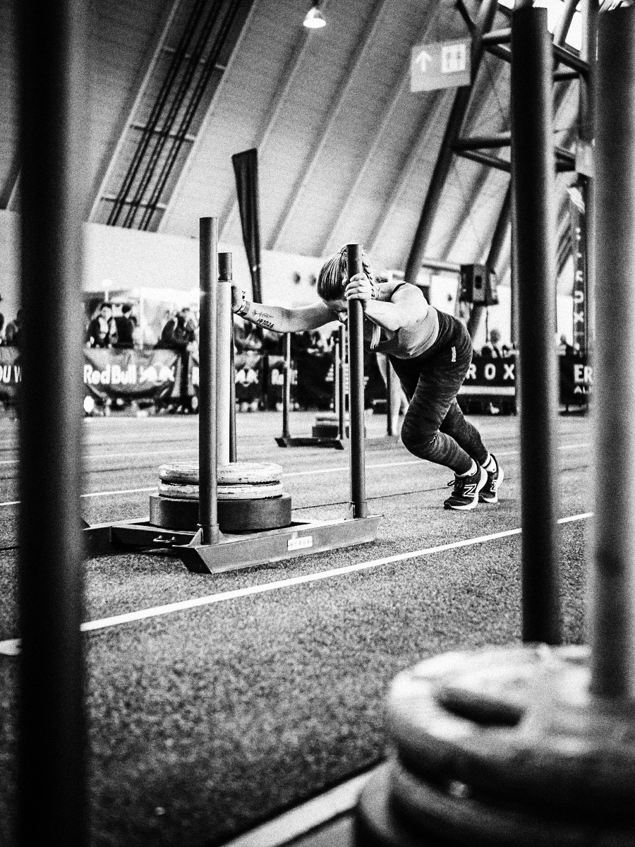 A woman pushing a weighted sled in an indoor athletic facility, participating in a strength training exercise, with spectators and banners visible in the background.